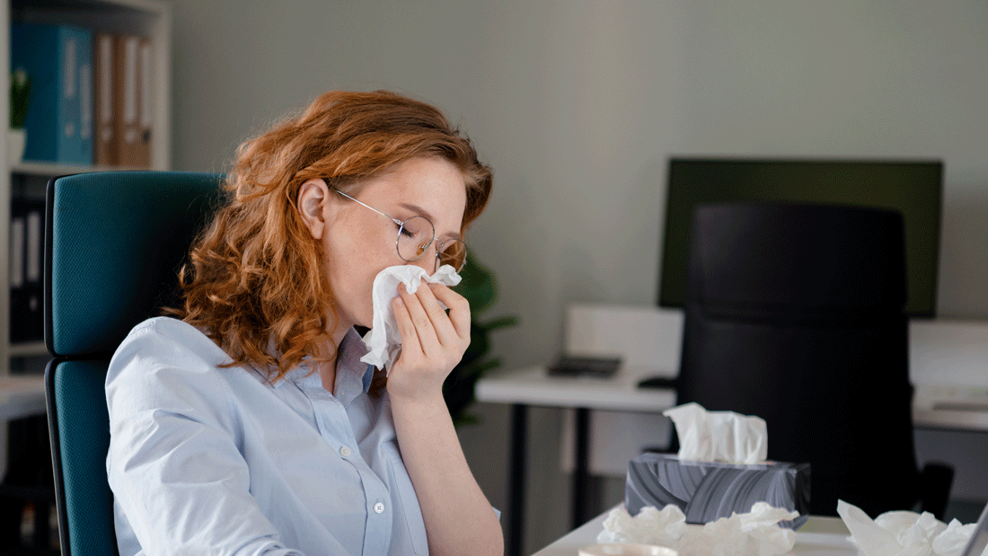 Das Bild zeigt eine Person in einem Büro, die eine Tasse hält und von Taschentüchern umgeben ist. Es deutet auf eine mögliche Erkrankung hin, wie eine Erkältung oder Grippe, während die Person dennoch am Arbeitsplatz ist. 