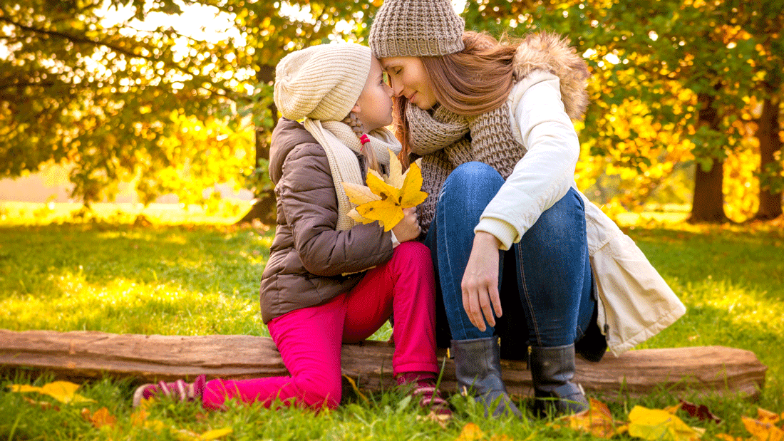 Das Bild zeigt eine herbstliche Szene mit zwei Personen (Mutter und Kind), die warm gekleidet sind und sich in einer natürlichen Umgebung befinden. Das Kind hält ein gelbes Herbstblatt, während die Mutter sich liebevoll zu ihm neigt.