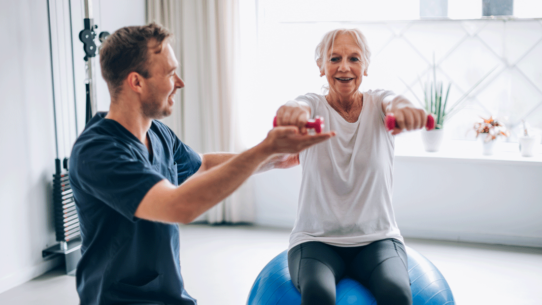 Eine ältere Frau trainiert mit kleinen Hanteln auf einem Gymnastikball, angeleitet von einem Therapeuten.