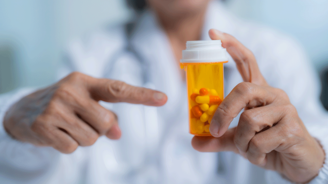 Dieses Bild zeigt eine Person, die eine Medikamentenflasche mit Kapseln oder Tabletten hält und darauf zeigt. Es scheint sich um eine medizinische Fachkraft, möglicherweise einen Arzt oder Apotheker, zu handeln, die auf das Medikament hinweist.