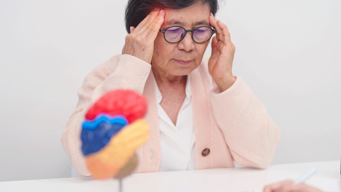 Das Bild zeigt eine Beratungssituation, in der eine Patientin möglicherweise über Beschwerden oder Schmerzen spricht. Auf dem Tisch steht ein Modell des menschlichen Gehirns, was darauf hindeutet, dass es sich um ein Gespräch über neurologische oder kognitive Gesundheit handeln könnte. 