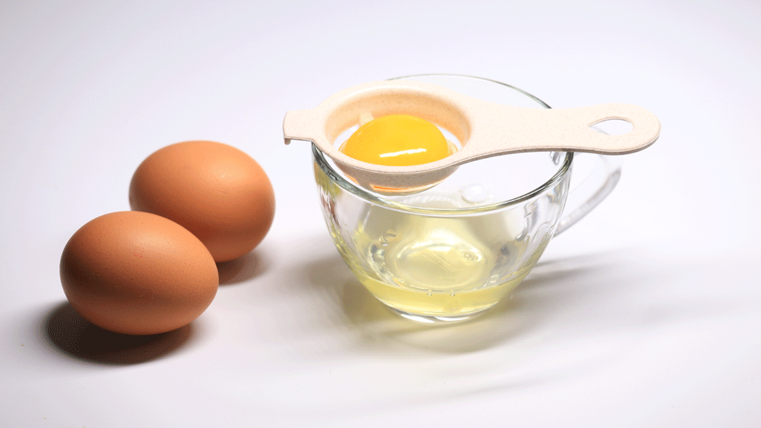 Das Bild zeigt zwei ganze Eier und ein aufgeschlagenes Ei, bei dem das Eigelb mithilfe eines Trennwerkzeugs vom Eiweiß getrennt wird. Das Eigelb befindet sich im Trennwerkzeug, während das Eiweiß in einem Glas darunter aufgefangen wird.