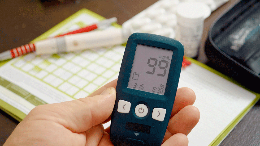 Das Bild zeigt ein Blutzuckermessgerät, das einen Wert von 99 mg/dL anzeigt. Im Hintergrund sind ein Kalender, ein Stift und Medikamente zu sehen, die auf das Management von Diabetes hinweisen. 
