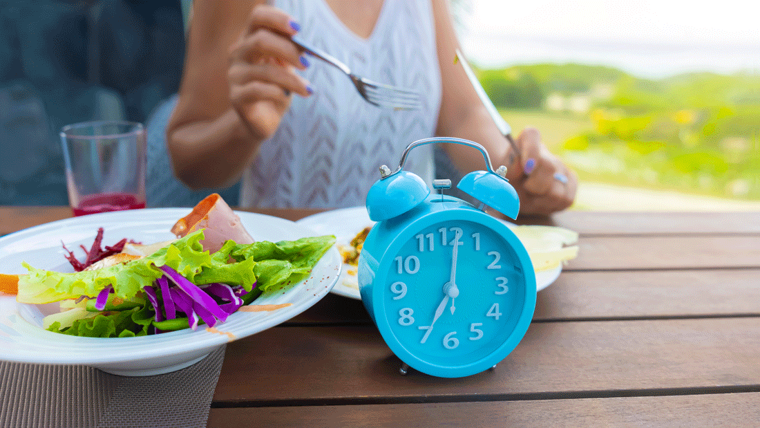 Das Bild zeigt eine Mahlzeit mit frischen Zutaten wie Salat und Gemüse, begleitet von einer blauen Uhr auf einem Tisch.