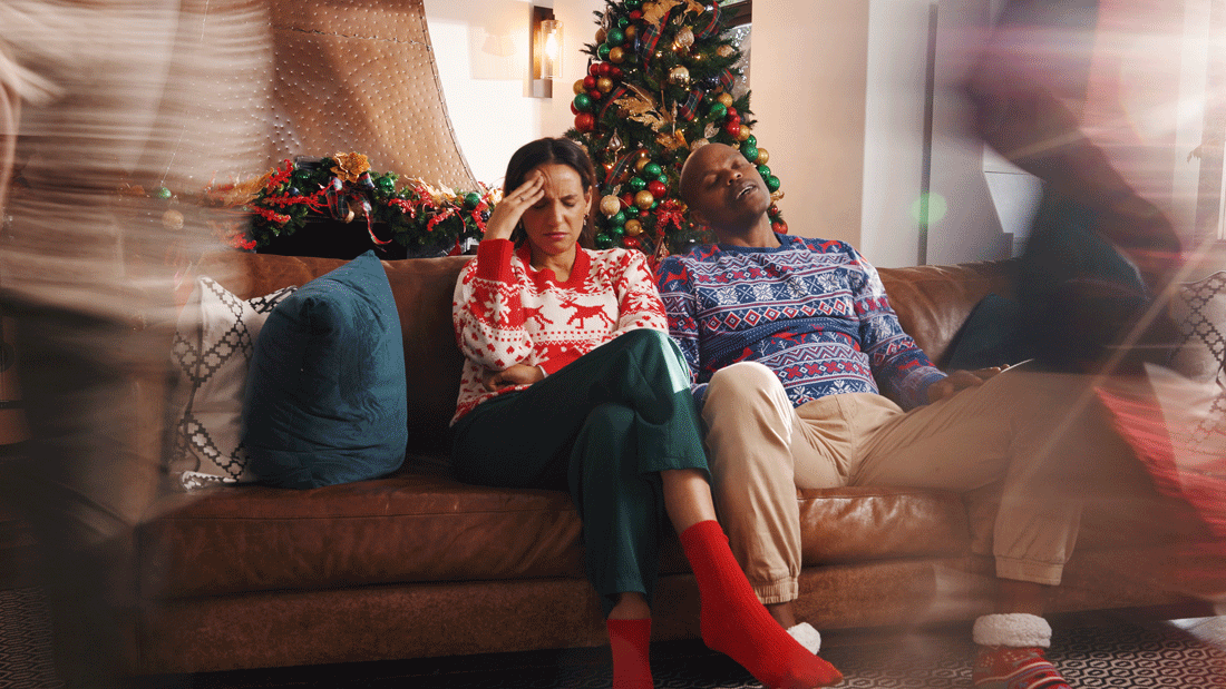 Das Bild zeigt eine Weihnachtsszene mit einem geschmückten Weihnachtsbaum im Hintergrund. Gestresste Personen sitzen auf einem Sofa.