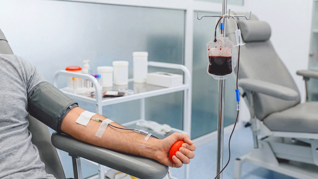 Eine Person spendet Blut und hält einen roten Ball in der Hand. Über einen Schlauch fließt Blut in einen Beutel am Infusionsständer; die Szene zeigt eine Blutspende in klinischer Umgebung.
