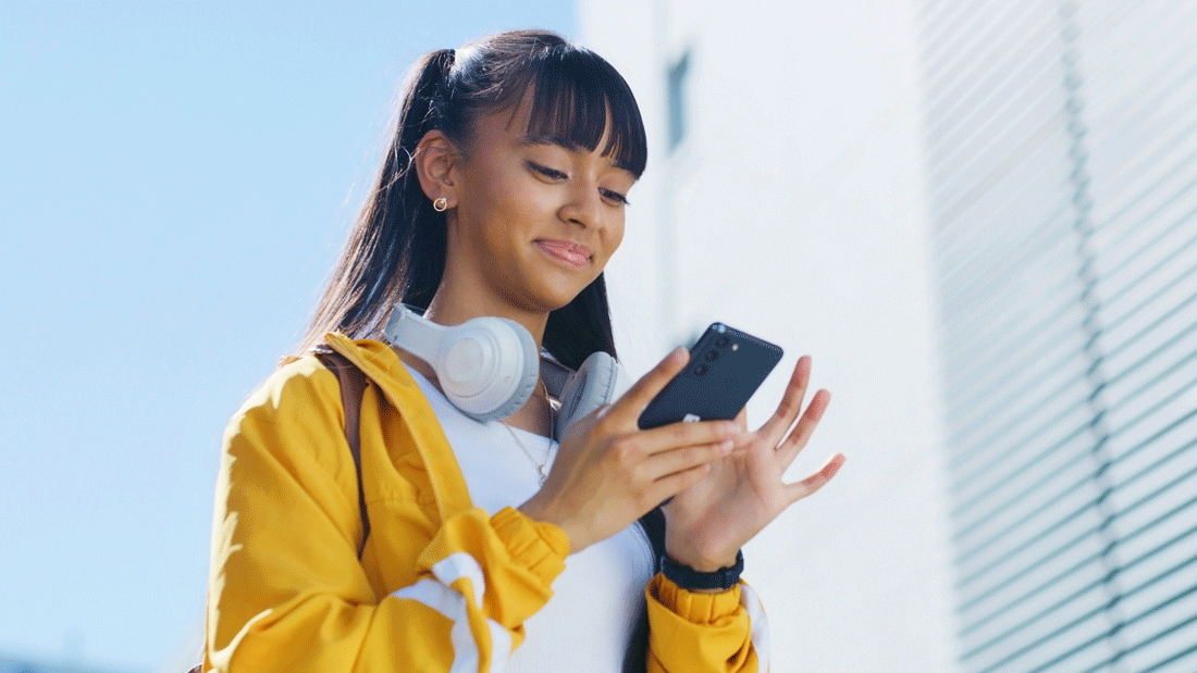 Dieses Bild zeigt eine Person, die ein Smartphone benutzt. Sie trägt eine gelbe Jacke und hat Kopfhörer um den Hals. Der Hintergrund zeigt eine moderne Architektur mit klaren Linien und einem blauen Himmel.