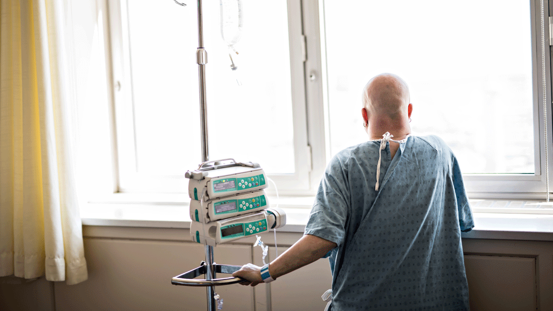 Kahlköpfige Person im Klinikhemd steht am Fenster neben Infusionsständer und Perfusoren.