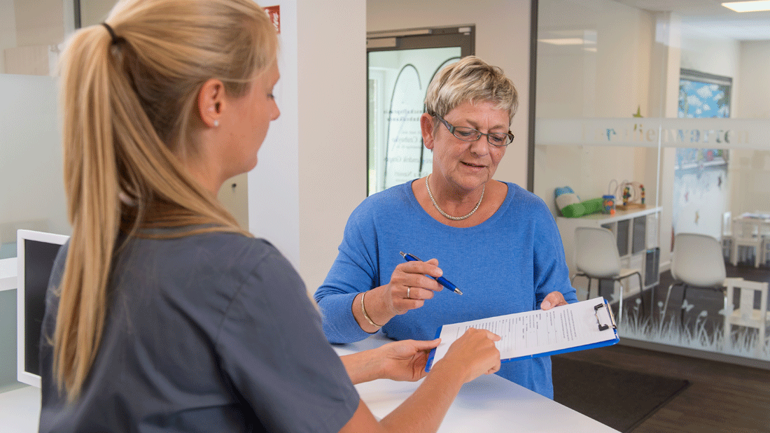 Das Bild zeigt eine Szene in einer medizinischen Einrichtung oder einem Empfangsbereich, in der eine Person ein Formular ausfüllt oder übergibt. Eine Mitarbeiterin in medizinischer Kleidung assistiert dabei. 
