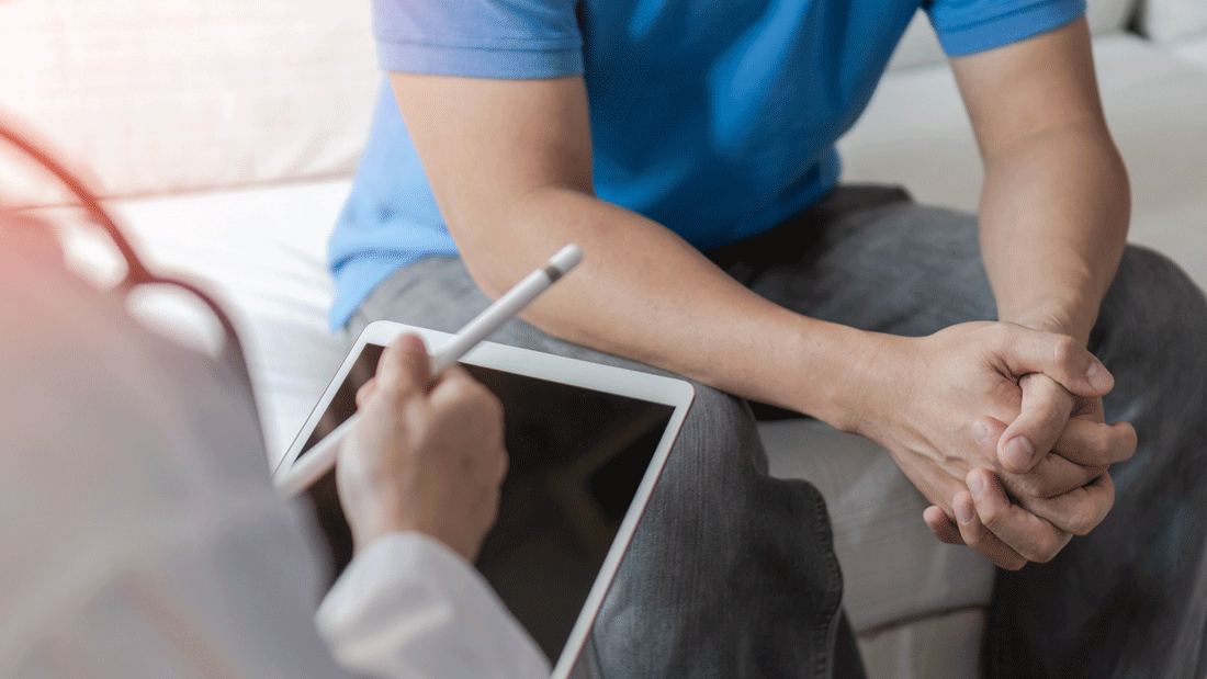 Ein Mann im blauen Shirt sitzt mit verschränkten Händen im Gespräch, gegenüber notiert medizinisches Personal etwas auf einem Tablet.