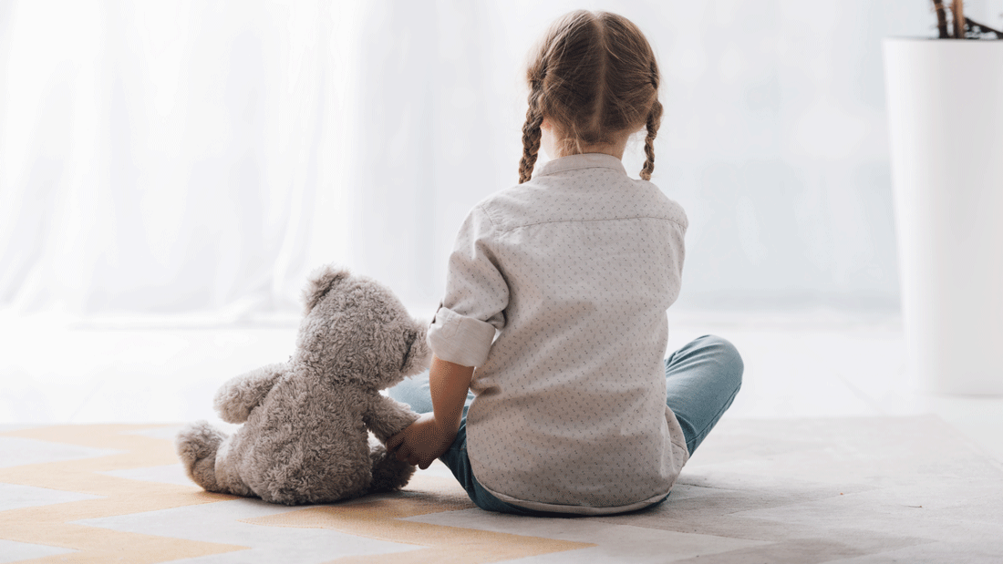 Ein kleines Kind sitzt mit dem Rücken zur Kamera auf dem Boden und hält einen Teddybären.