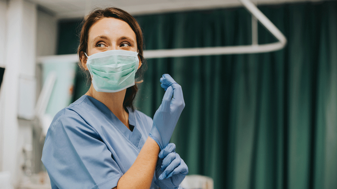 Das Bild zeigt eine medizinische Fachkraft in einer klinischen Umgebung, die Schutzkleidung und Handschuhe trägt und sich auf eine Untersuchung oder einen Eingriff vorbereitet. 
