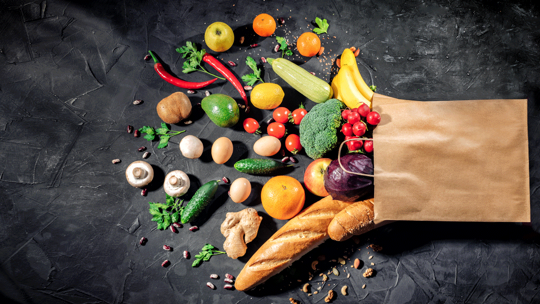 Das Bild zeigt eine Vielzahl von frischen Lebensmitteln, darunter Obst, Gemüse, Eier, Brot und Nüsse, die aus einer Papiertüte herausragen.