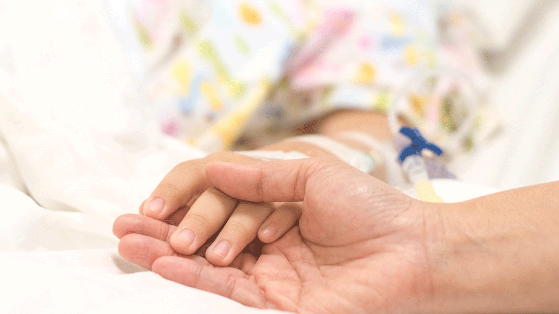 Eine erwachsene Hand hält behutsam die Hand eines Kindes im Krankenhausbett. Ein Venenzugang ist sichtbar.