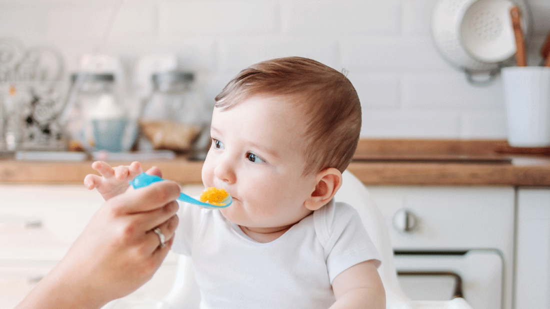 Das Bild zeigt eine Szene, in der ein Baby in einem Hochstuhl gefüttert wird. 