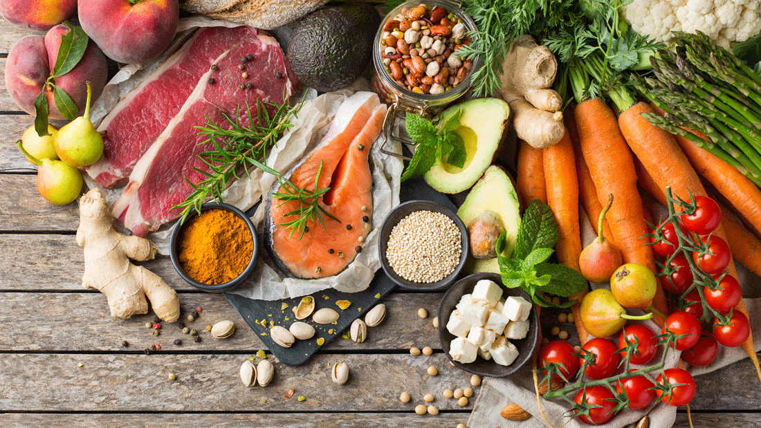 Das Bild zeigt eine Vielzahl von frischen und gesunden Lebensmitteln, darunter Obst (Pfirsiche, Birnen, Avocado), Gemüse (Karotten, Tomaten, Blumenkohl, Spargel), Proteine (Lachs, Fleisch, Käse), Hülsenfrüchte, Nüsse, Gewürze (wie Kurkuma) und Vollkornbrot. Es symbolisiert eine ausgewogene Ernährung, die reich an Nährstoffen ist und die Grundlage für einen gesunden Lebensstil bildet.