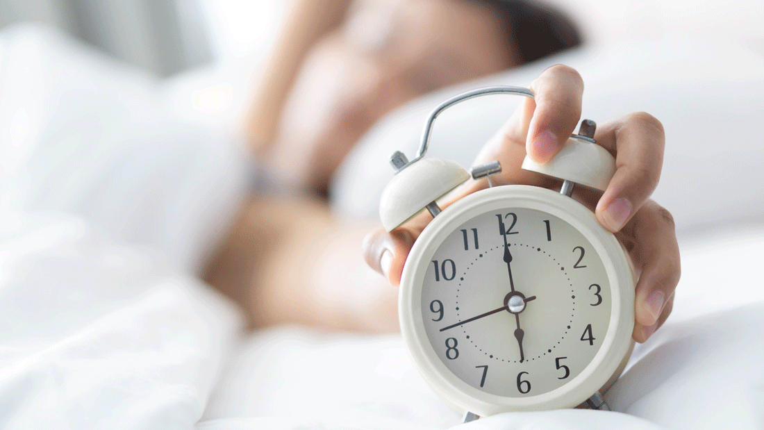 Unscharf im Bett liegende Person greift nach einem weißen Wecker.