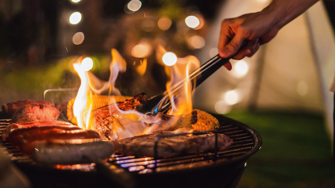  Nächtliche Grillszene: Eine Hand wendet mit der Zange Fleisch und Mais über offenen Flammen; Würstchen liegen auf dem heißen Rost.