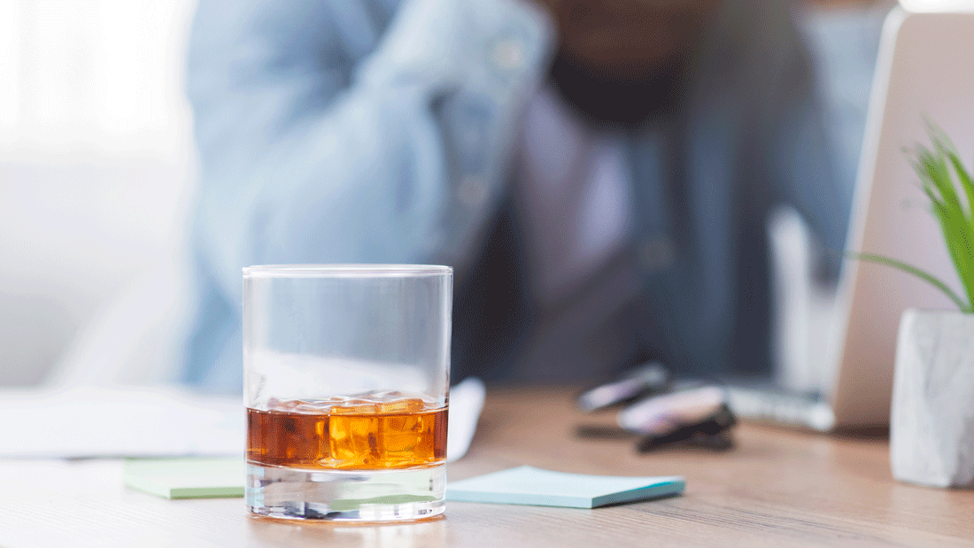 Dieses Bild zeigt ein Glas mit einem alkoholischen Getränk, das auf einem Schreibtisch steht. Im Hintergrund ist eine Person unscharf zu erkennen, die möglicherweise gestresst oder nachdenklich wirkt.
