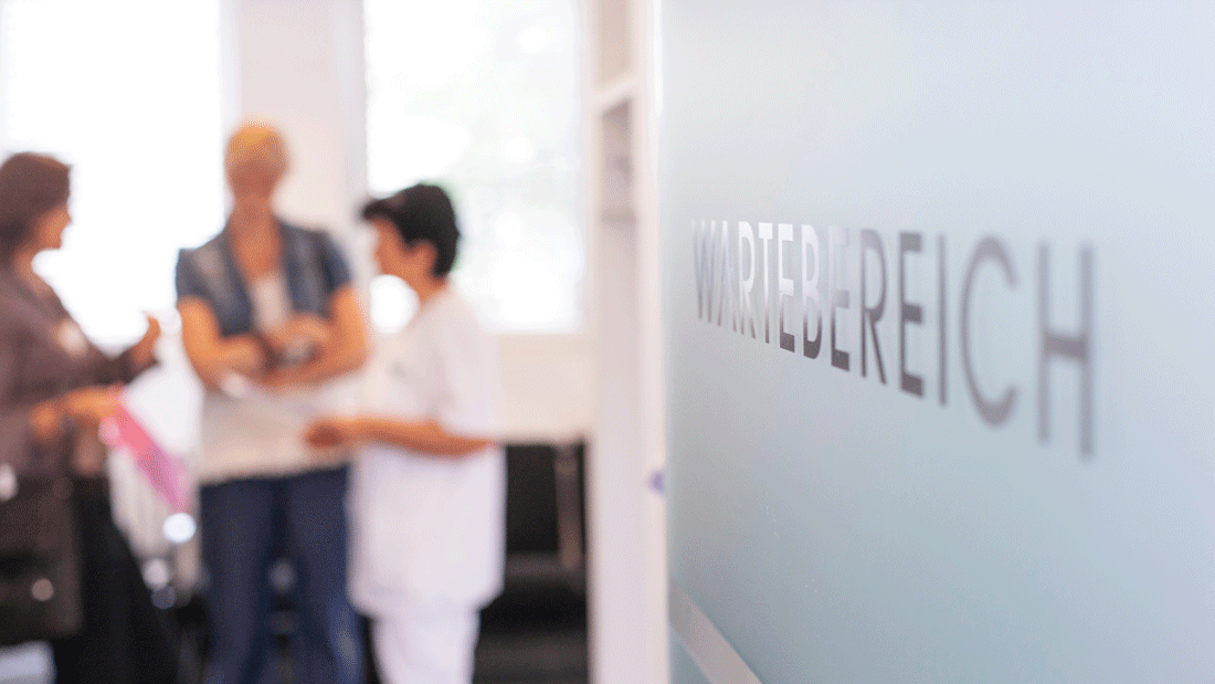 Das Bild zeigt einen Wartebereich, der durch eine Glastür oder -wand gekennzeichnet ist. Im Hintergrund sind Personen in einer Gesprächssituation zu sehen, möglicherweise in einer medizinischen Einrichtung oder Praxis. 