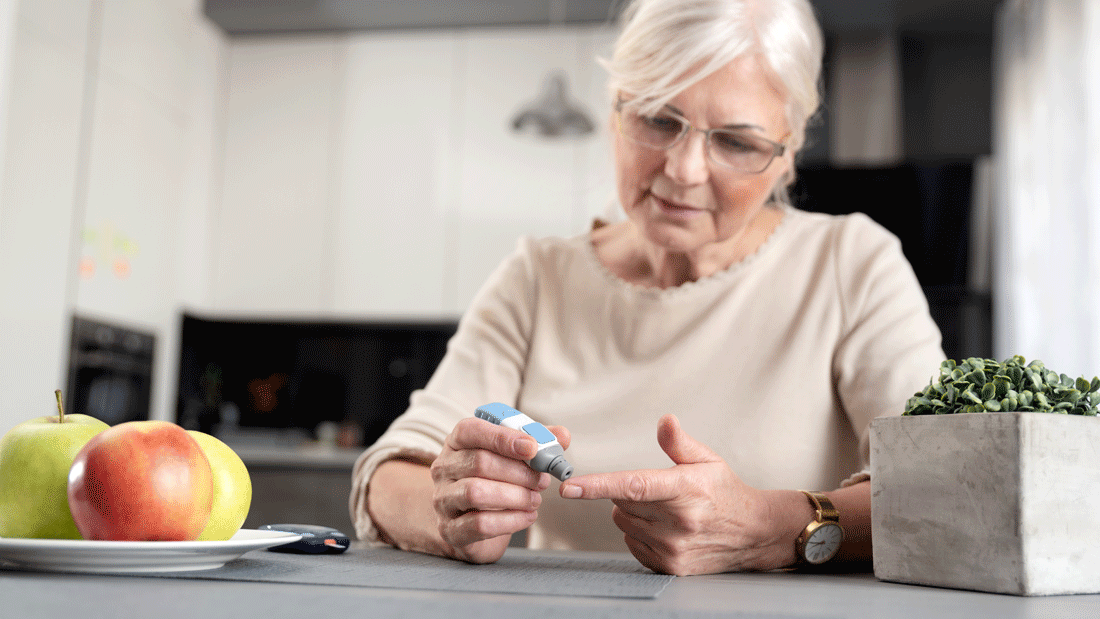 Das Bild zeigt eine ältere Frau, die ihren Blutzucker misst. Die Äpfel auf dem Tisch könnten auf eine ausgewogene Ernährung hinweisen, die für Diabetiker von großer Bedeutung ist.
