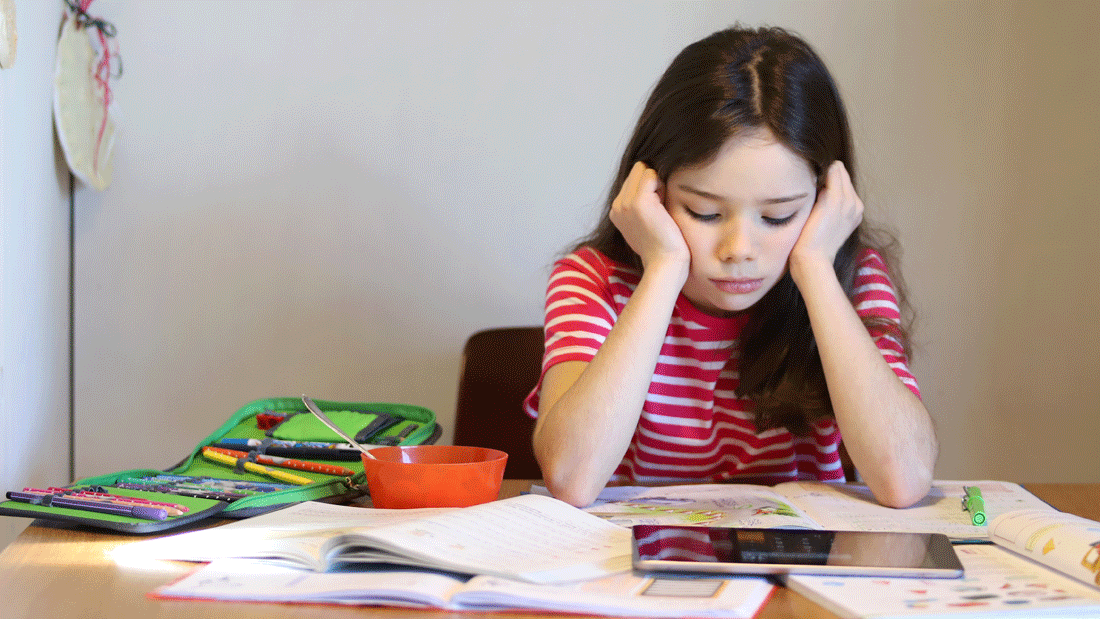 Ein Mädchen sitzt nachdenklich am Tisch vor Heften, Büchern und Tablet. Neben Schulmaterial steht eine Schüssel.