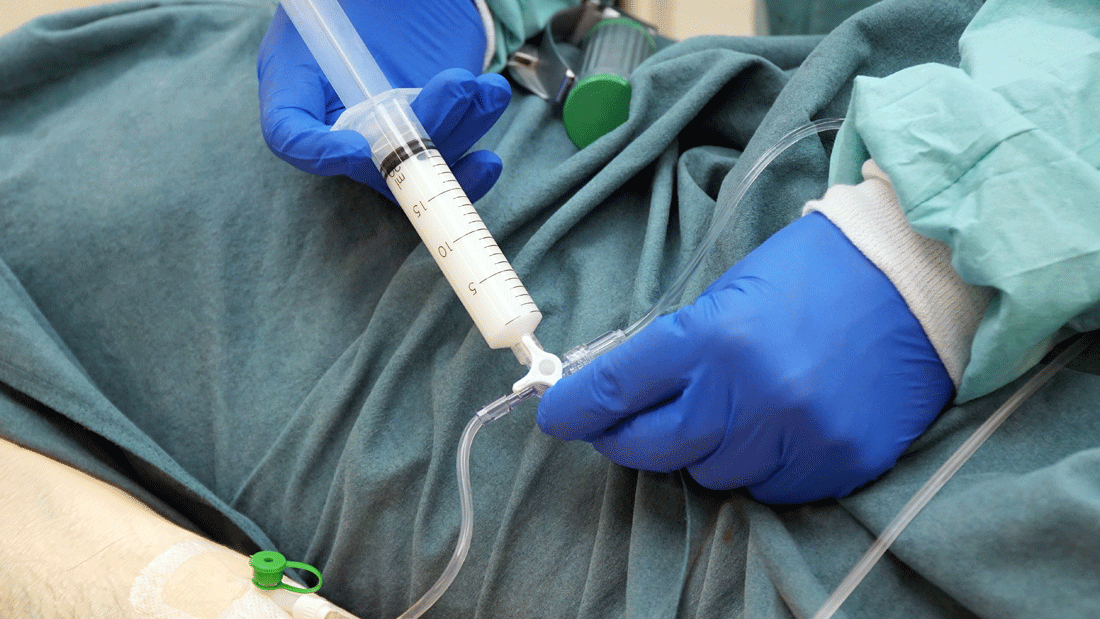 Arzt in Schutzkleidung hält eine Spritze an einen Schlauchzugang am liegenden Patienten.