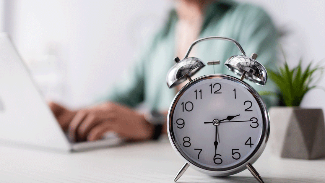 AdobeStock_366742567.gif Dieses Bild zeigt einen klassischen Wecker im Vordergrund, der die Bedeutung von Zeitmanagement und Pünktlichkeit symbolisiert. Im Hintergrund ist eine Person zu sehen, die an einem Laptop arbeitet.