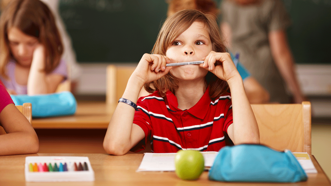 Dieses Bild zeigt eine Schulszene, in der ein Kind an einem Tisch sitzt, umgeben von Schulmaterialien wie Buntstiften und einer Brotdose. Im Hintergrund sind weitere Kinder und eine Tafel zu sehen. 