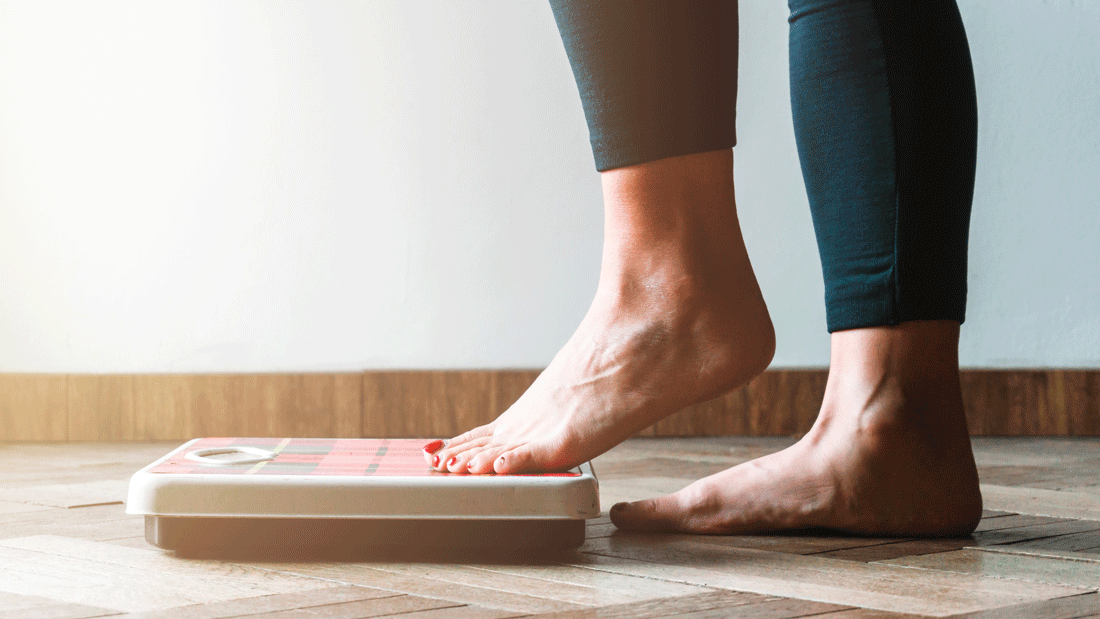  Dieses Bild zeigt eine Person, die auf eine Waage tritt, vermutlich um ihr Gewicht zu messen. Der Fokus liegt auf den Füßen und der Waage, mit einem schlichten und minimalistischen Hintergrund. 