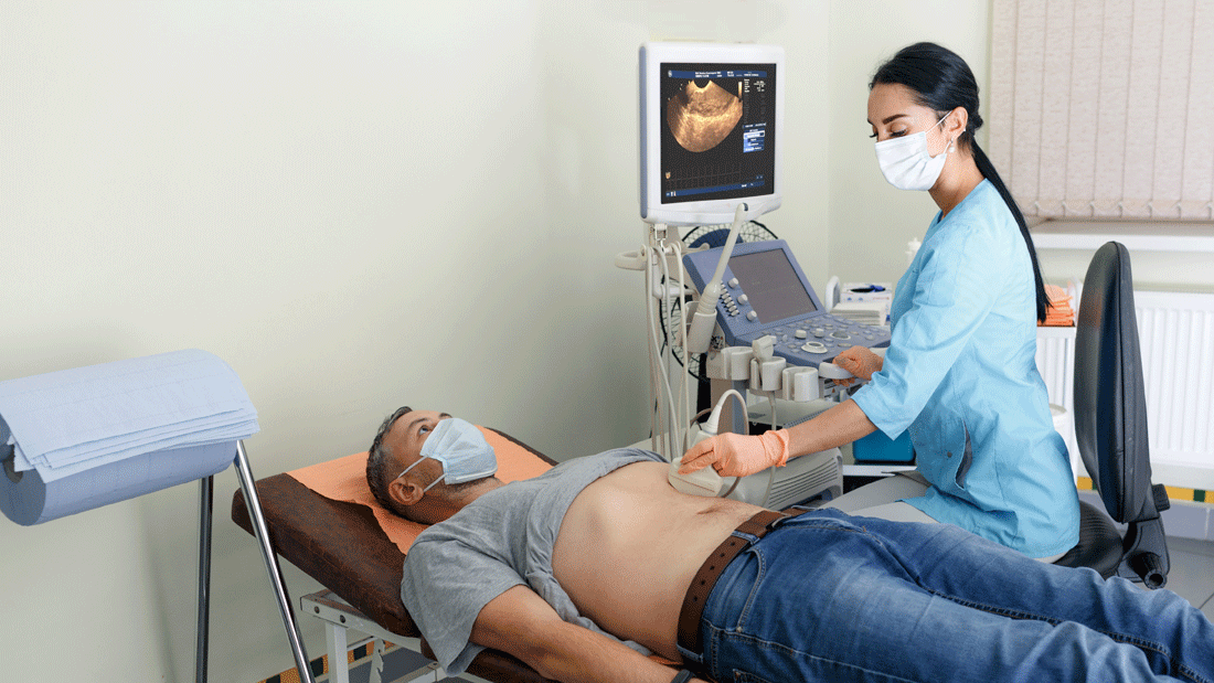Ein Mann liegt mit freigemachtem Bauch auf der Untersuchungsliege, während eine Fachkraft per Ultraschall den Bauchraum untersucht. Beide tragen Masken; das Gerät zeigt das Sonografiebild.