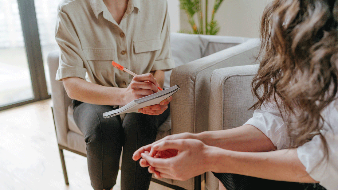 Das Bild zeigt eine Beratungssituation oder ein Gespräch zwischen zwei Personen. Eine Person hält einen Notizblock und einen Stift, während sie scheinbar Notizen macht. Die andere Person sitzt gegenüber und scheint in das Gespräch vertieft zu sein. 