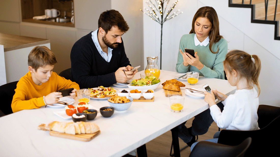 Dieses Bild zeigt eine Familie, die gemeinsam am Esstisch sitzt. Auf dem Tisch stehen verschiedene Speisen wie Brot, Croissants, Salat und kleine Schalen mit Snacks. Alle Personen sind mit ihren Smartphones beschäftigt, anstatt miteinander zu interagieren.