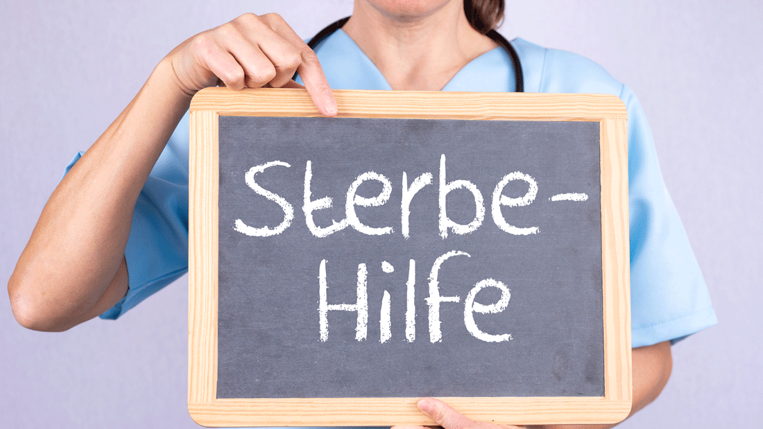 Eine Person in medizinischer Kleidung hält eine Tafel mit der Aufschrift „Sterbe-Hilfe“.
