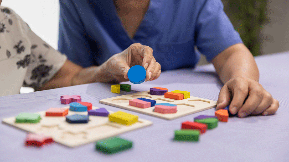 AdobeStock_536080275.gif Eine ältere Person und eine Pflege- oder Betreuungskraft in blauer Kleidung legen bunte Holzformen in Steckbretter.