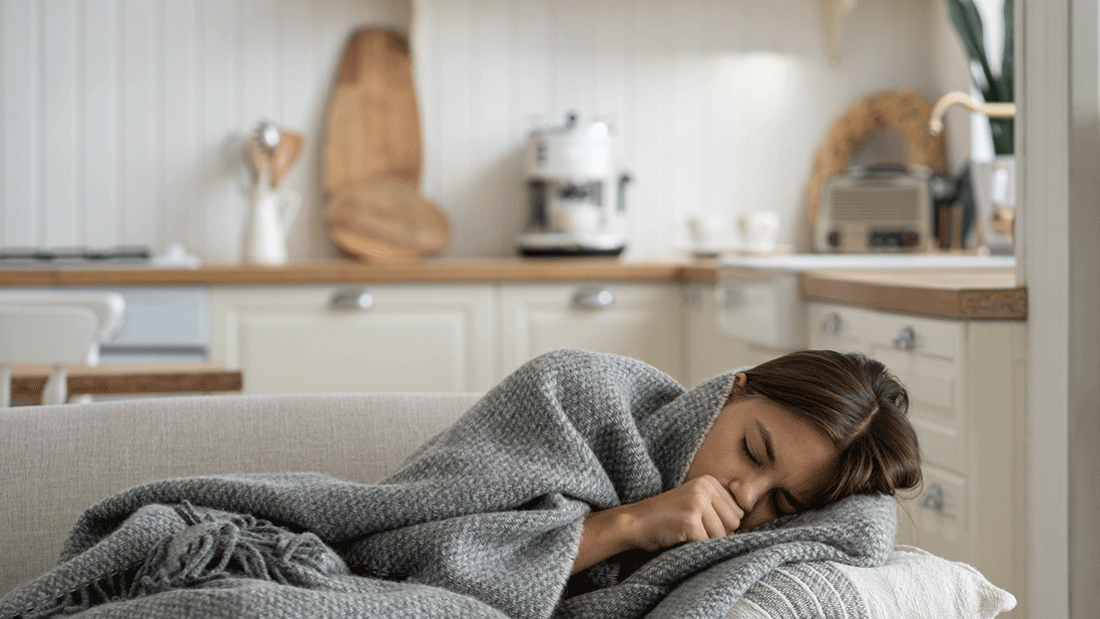 Das Bild zeigt ein möglicherweise an Grippe erkranktes Kind, das auf einem Sofa liegt und sich in eine warme, graue Decke eingekuschelt hat.