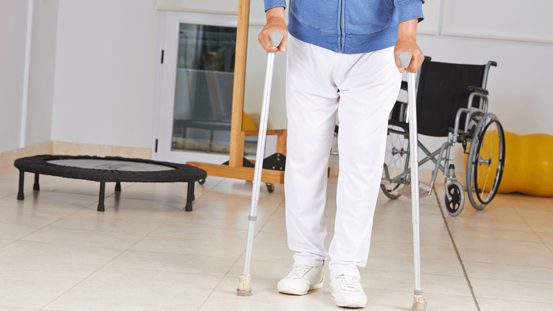 Dieses Bild zeigt eine Person, die mit Hilfe von Unterarmgehstützen geht. Im Hintergrund sind ein Rollstuhl, ein Trampolin und andere Gegenstände zu sehen, die auf eine physiotherapeutische oder rehabilitative Umgebung hinweisen.