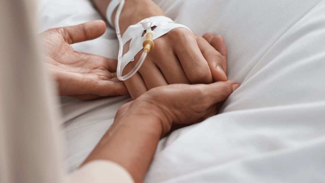 Dieses Bild zeigt eine Hand, die mit einem intravenösen Zugang verbunden ist, während eine andere Hand sie sanft hält. 