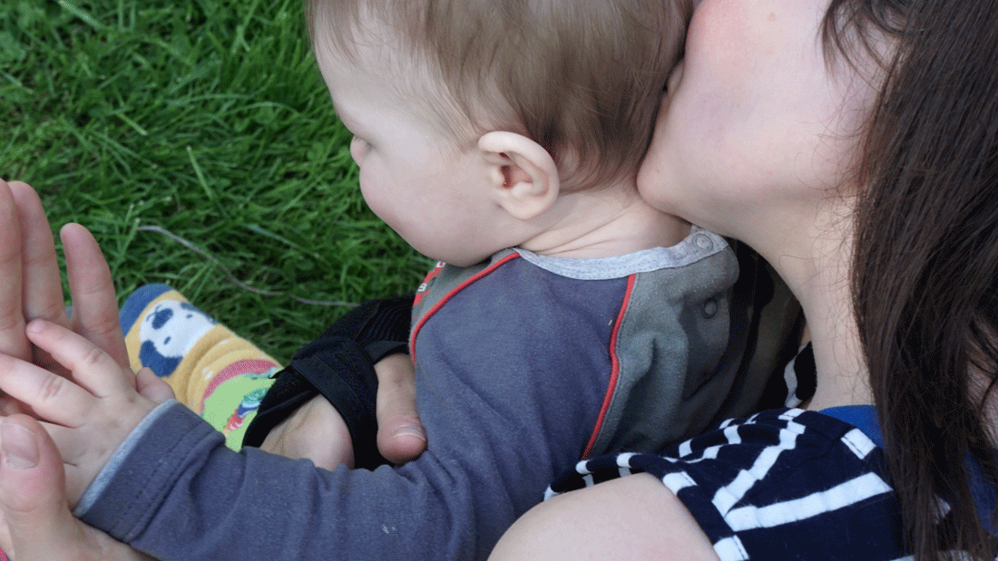 Eine Frau hält ein Baby im Arm und küsst es am Kopf. Beide sitzen im Gras.